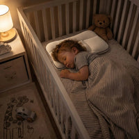 Toddler asleep on the pillow in a white crib at night, with a teddy bear and a lit bedside lamp.